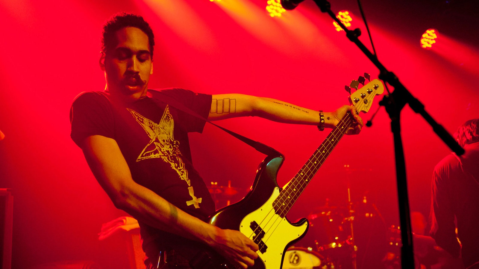 Bassist mit dunkelm Oberlippenbart spielt mit gestreckten Armen und geschlossenen Augen im roten Bühnenlicht.