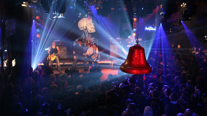 Blick von schräng oben auf die eine beleuchtete Bühne mit der Band Dirty Deeds 79'. Über dem Zuschauerraum hängt eine große Glocke.