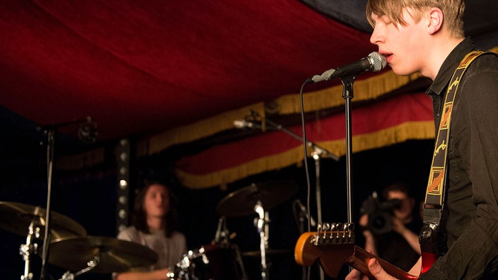 Aufnahme von Schlagzeuger im Hintergrund und  Sänger mit Mikrofon und Gitarre