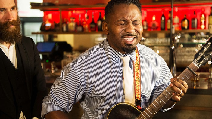 Mann mit verbissenem Gesicht beim Gitarre spielen, hinter ihm steht ein Mann mit Vollbart