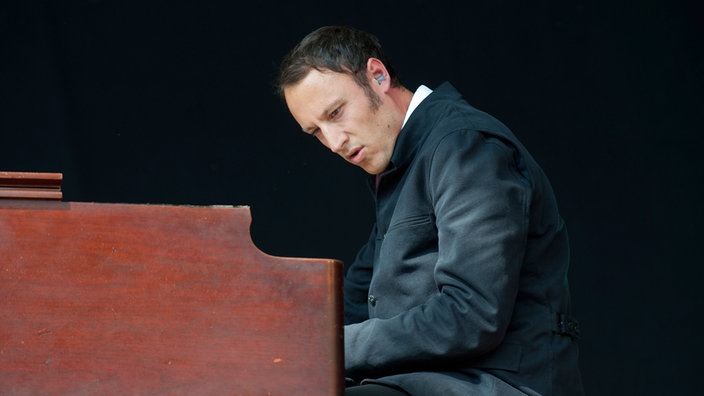 Mann im schwarzen Mantel sitzt an einer Orgel und schaut nach unten