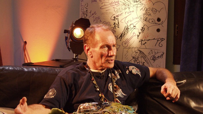 Mitglied der Band "Guru Guru" sitzt auf einem Sofa und erzählt etwas.