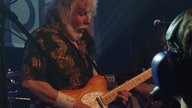 Mann mit glauen Haaren und gemustertem Hemd spielt Gitarre und schaut nach unten auf seine Hände.
