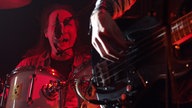 Singender Schlagzeuger im Hintergrund, Hand am Gitarrenkorpus im Vordergrund.