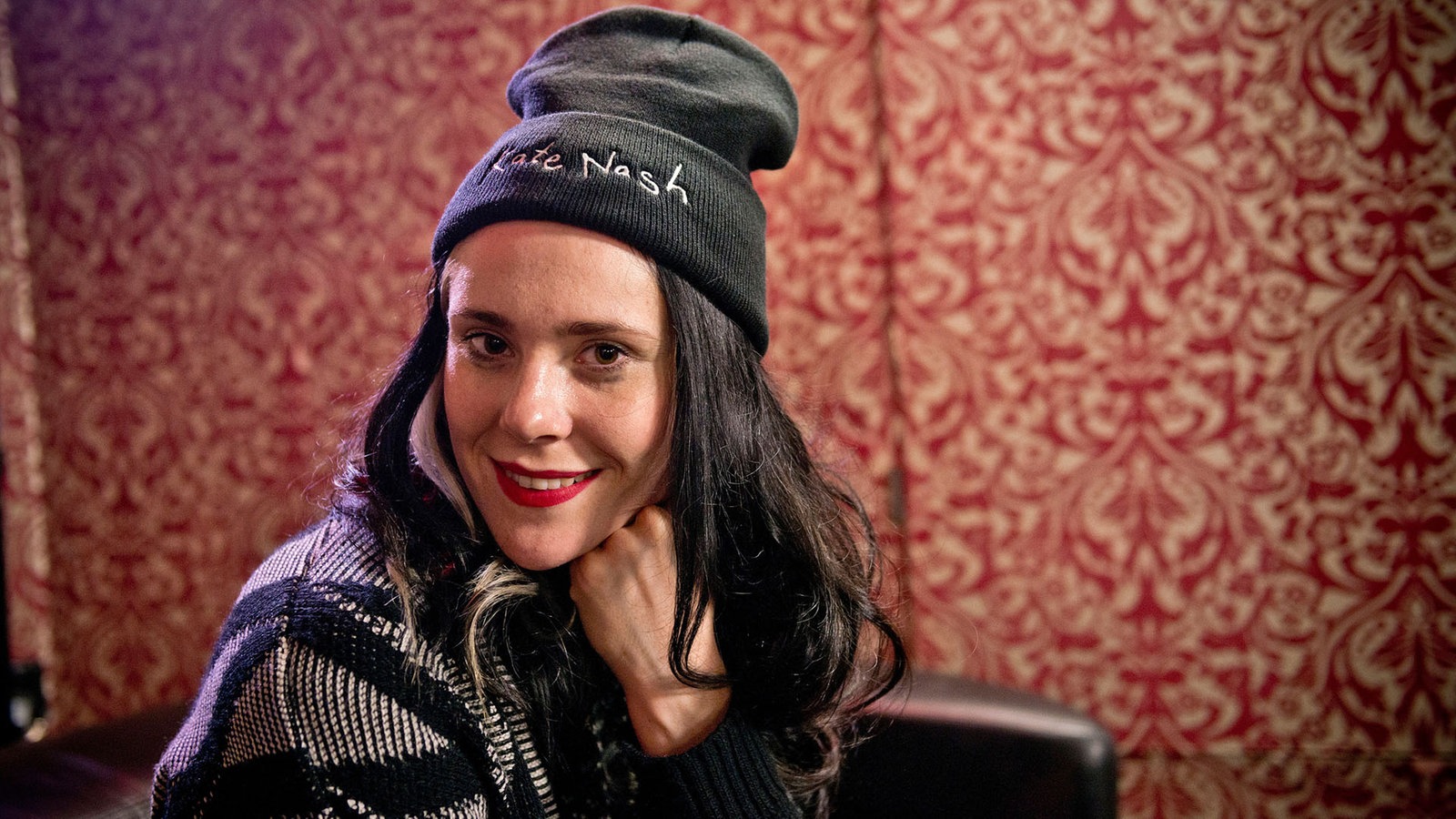 Kate Nash sitzt auf einer Couch die in einem Zimmer mit einer wilden rot-beigen Tapete steht.