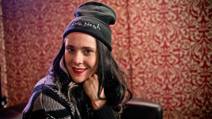 Kate Nash sitzt auf einer Couch die in einem Zimmer mit einer wilden rot-beigen Tapete steht.