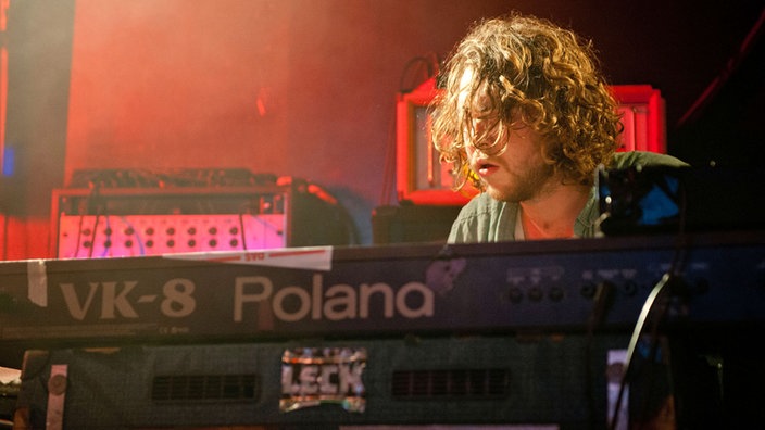Hinter seinem Keyboard guckt eines der Bandmitglieder gerade so hervor, während er mit dem spielen beschäftigt ist.