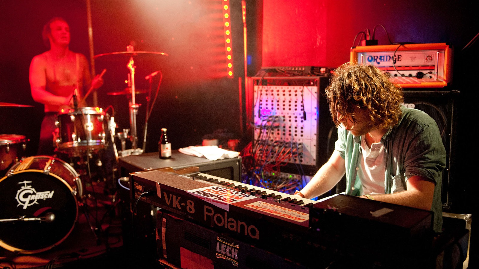 Im Vordergrund sitzt der weiß angestrahlte Keyboarder der Band während man im Hintergrund den ebenfalls oberkörperfreien Schlagzeuger sehen kann.