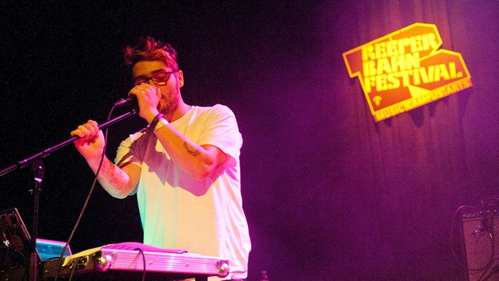 Der Keyboarder und Backing-Sänger der Band Breton steht vor seinem Keyboard am Mikrofon und singt mit geschlossenen Augen hinein.