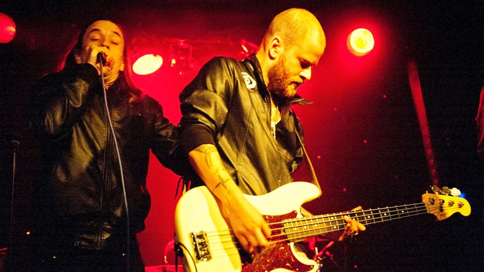 Ein kurzrasierter Bassist spielt, daneben der Sänger.