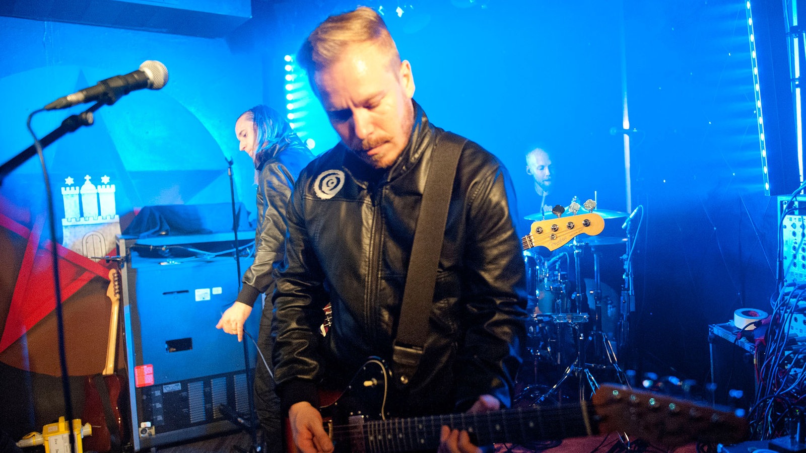 Der Gitarrist, der seinen Blick senkt und spielt. Er trägt eine blonde EBM-Tolle und eine schwarze Lederjacke.