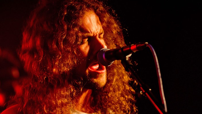 Der langhaarige Bassist singt mit geschlossenen Augen in das Mikrofon, welches rot angestrahlt wird.