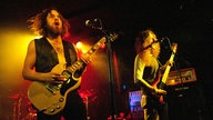 Der GItarrist und der Bassist der Band stehen im Vordergrund und spielen exzessiv, während im Hintergrund das Schlagzeug rosa rot angestrahlt wird.
