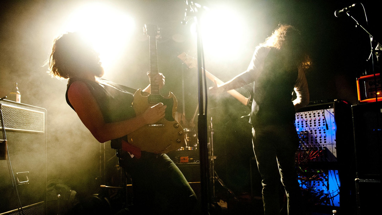 Der Gitarrist und der Bassist der Band stehen beide vor gleizend weißem Licht in Dunst der Bühne und spielen auf ihren Instrumenten.