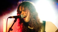 Der blau-rot angestrehlte Bassist hat die Haare ins Gesicht hängen und singt mit weit offenem Mund in das Mikrofon.