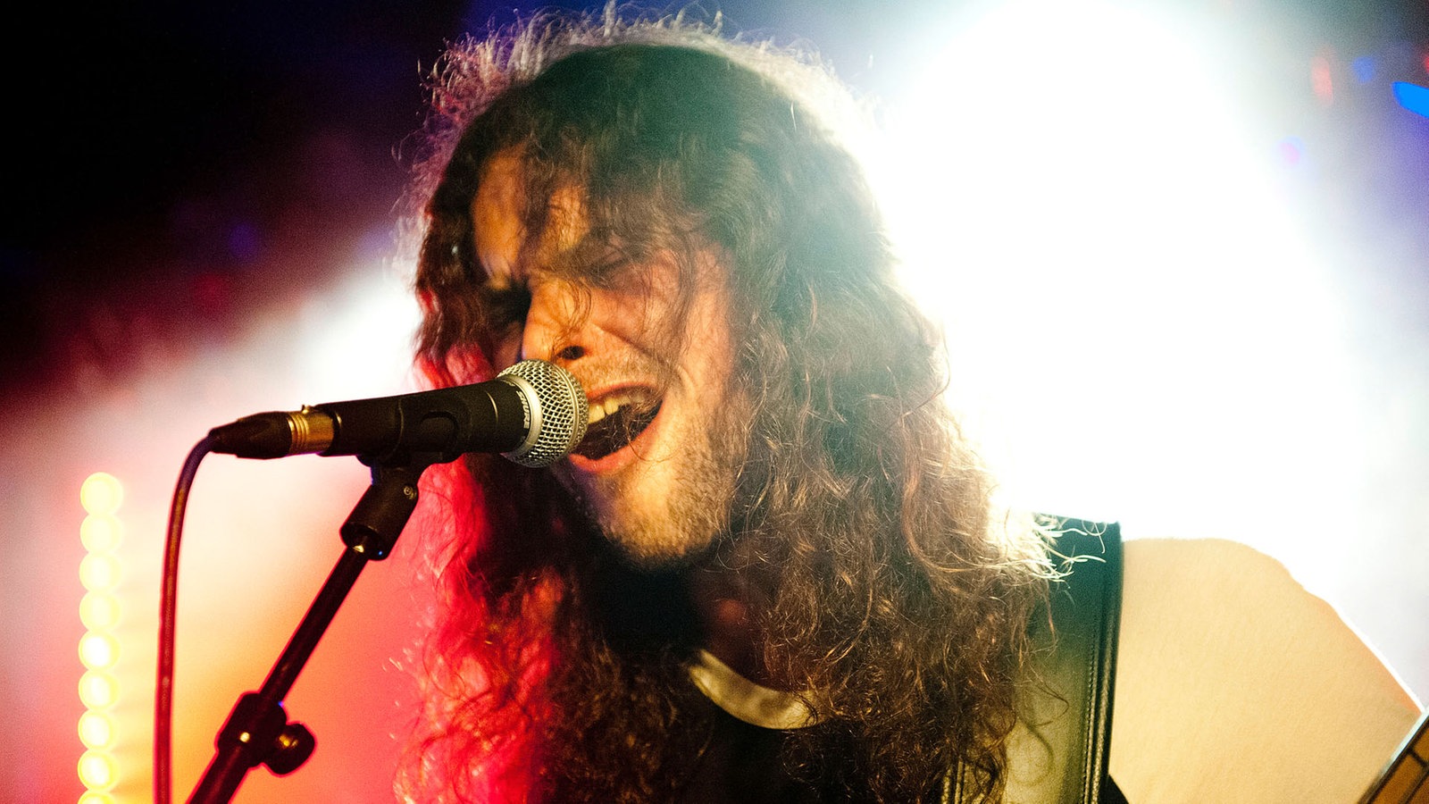 Der blau-rot angestrehlte Bassist hat die Haare ins Gesicht hängen und singt mit weit offenem Mund in das Mikrofon.