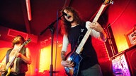 Die Beiden Männer mit den Saiteninstrumenten sind aus dem Publikum heraus zu sehen , während der Sänger der Band in das Mikrofon zu schreien scheint.