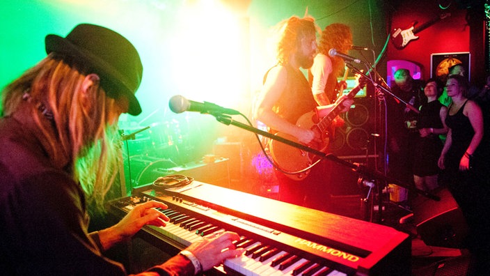 Im Vordergrund ist der Keyboarder zu sehen welcher mit nach unten geneigtem Kopf spielt, während im Hintergrund der Rest der Band in grünem Licht erstrahlt.