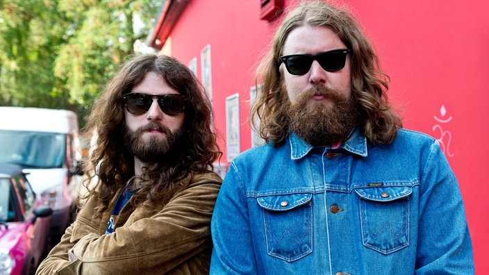 Zwei Bandmitglieder der Band "The Sheepdogs" stehen vor einer roten Wand. Beide haben einen langen Bart, lange Haare und tragen dunkle Sonnenbrillen.