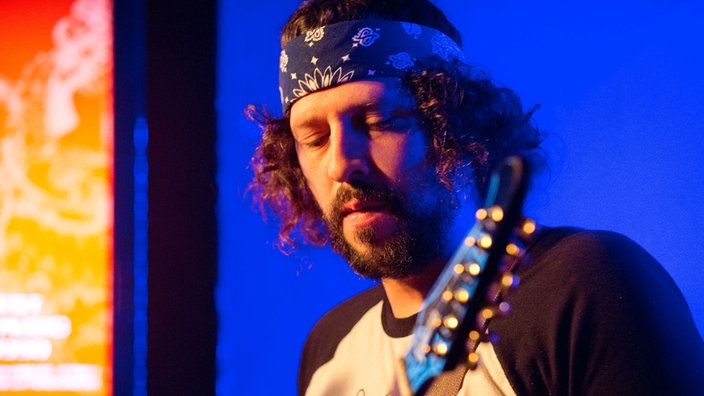 Einer der Gitarristen hat ein dunkles Bandana um seine Stirn gewickelt und sieht angestrengt nach unten auf seine Gitarre.