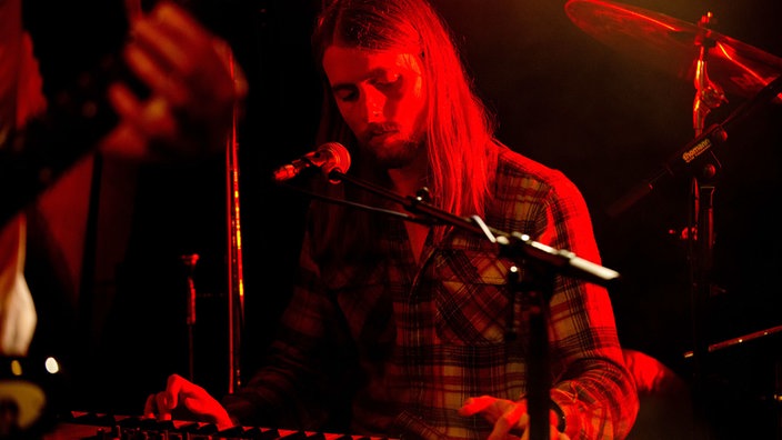 Der Keyboarder der Band sieht etwas gelangweilt nach unten auf sein Instrument und wird dabei rot von der Seite angestrahlt.