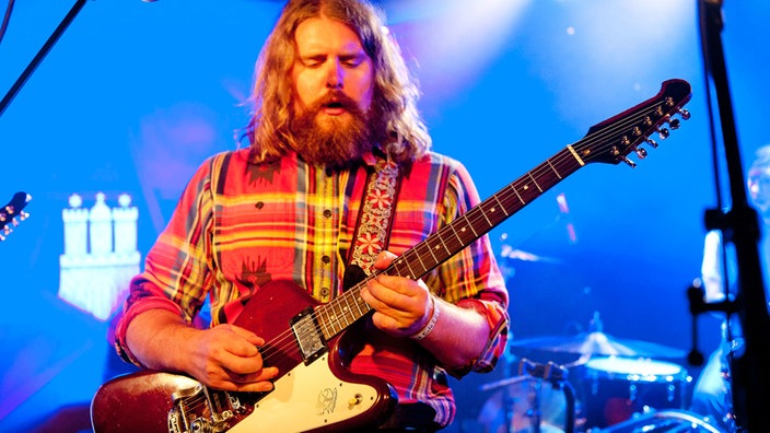 Der rundliche Frontmann der Band sieht angestrengt nach unten und spielt auf seiner Gitarre.