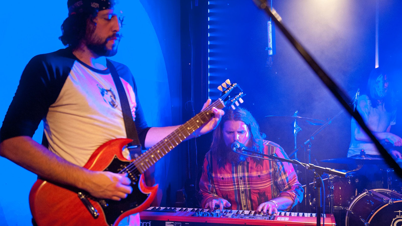 Ein weiters Bandmitglied steht mit einer Gitarre im Vordergrund währen im Hintergrund der Keyboarder am Mikrofon mitsingt.