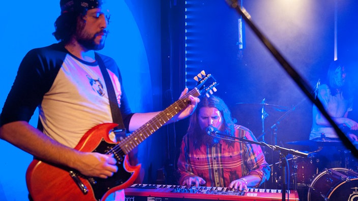 Ein weiters Bandmitglied steht mit einer Gitarre im Vordergrund währen im Hintergrund der Keyboarder am Mikrofon mitsingt.