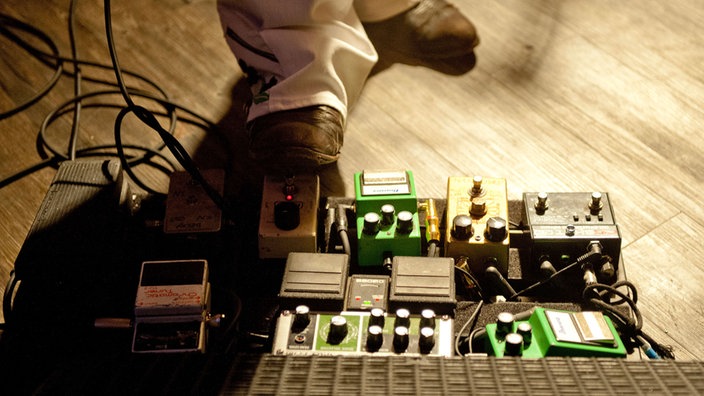 Man sieht den Fuß einer der Gitarristen, der gerade eines seiner Effektgeräte für die Gitarre am bedienen ist.