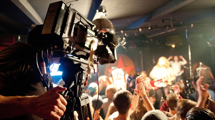 Im Vordergrund ist eine der Filkameras des Rockpalast zu sehen während im Hintergrund die Menge die Band abfeiert.