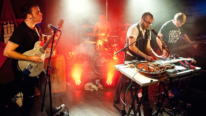Aktionsreiche Aufnahme aller Antlered Man Musiker auf der Bühne: hinten der Drummer, davor Bassist, Gitarrist und der Effekte spielende Multiinstrumentalist - alle in Aktion.