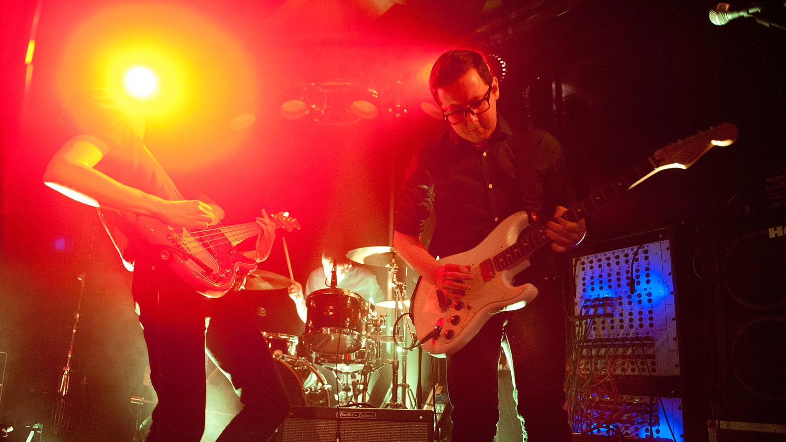 Im Hintergrund der Schlagzeuger, davor Bassist und Gitarrist der Band. Ausserdem ist ein Geräte-Rack zu sehen, an dem die vielen Kabel der Instrumente angeschlossen sind.