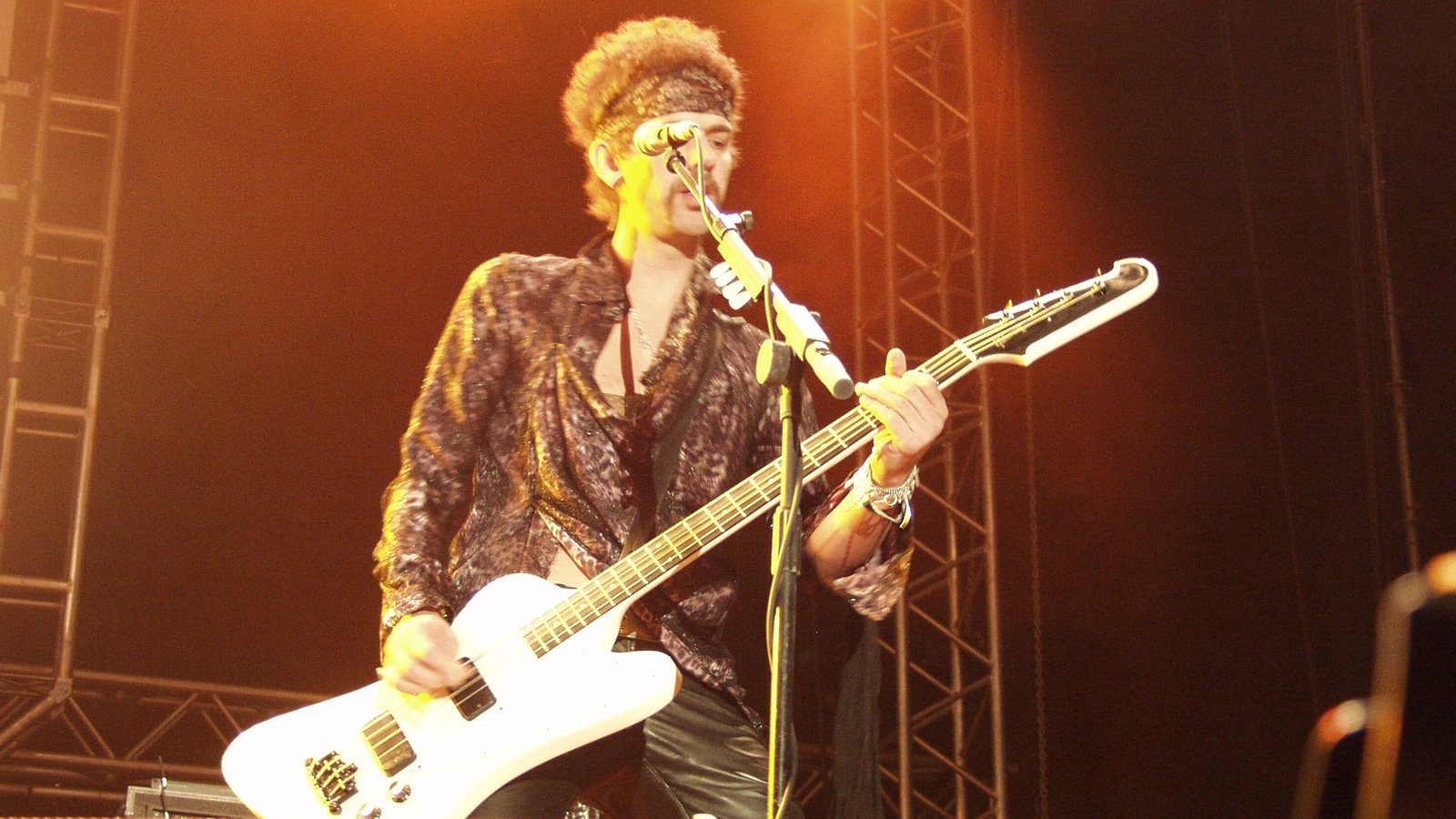 Mann mit Glitzerhemd und Stirnband spielt Bass.