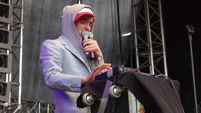 Winson mit Kaputzenpullover und zwei Mikrofonen in der Hand steht an einem Keyboard.