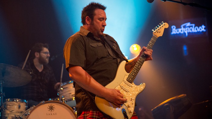 Gitarrist von King King spielt ein heißes Solo auf seiner weißen Fender Stratocaster Gitarre, im Hintergrund ist das Rockpalast Logo zu sehen