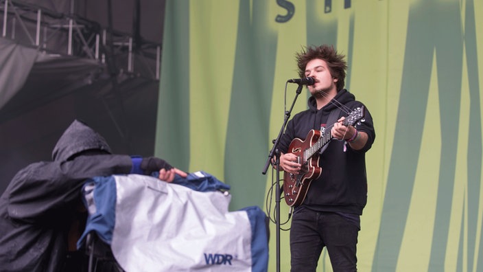 Der Gitarre spielende und singende Clemens wird von einem in Regenmantel gehüllten Kameramenschen gefilmt. Auch die Kamera ist regensicher verpackt.