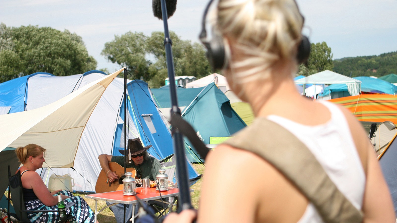 Junge Tonfrau mit Tonangel vorne, weiter hinten unterm Vorzelt zwei Festivalbesucher die ihr Gitarrenspiel genießen