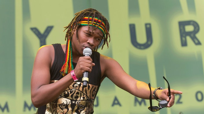Der Sänger von Raging Fyah hält die Sonnenbrille in der Hand, zeigt auf jemandem und singt diesem jemandem zu.