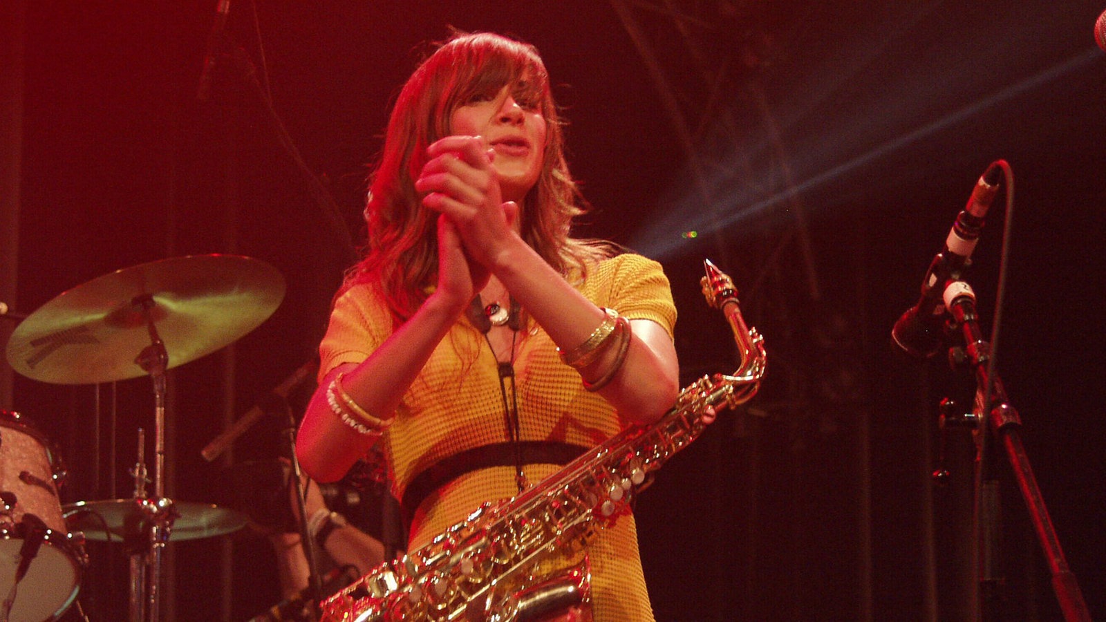 The Zutons: Eine Frau im gelben Kleid hat ein Saxophon umgehängt und klatscht im roten Bühnenlicht