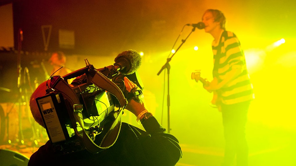 Im Vordergrund ist ein Kameramann des Rockpalasts zu sehen und im Hintergrund in gelbes Licht getaucht der Sänger der Band An Horse.
