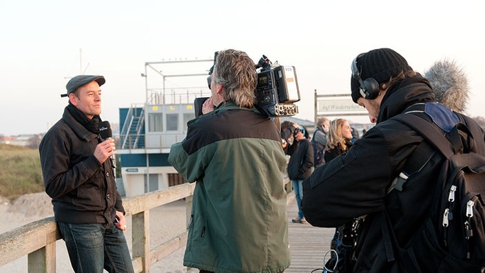 Manuel Unger lehnt an einem Geländer am Strand und wird von dem Lamerateam gefilmt.