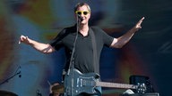Mann mit gelber Sonnenbrille, umgehängter Gitarre und schwarzem T-Shirt breitet seine Arme aus.