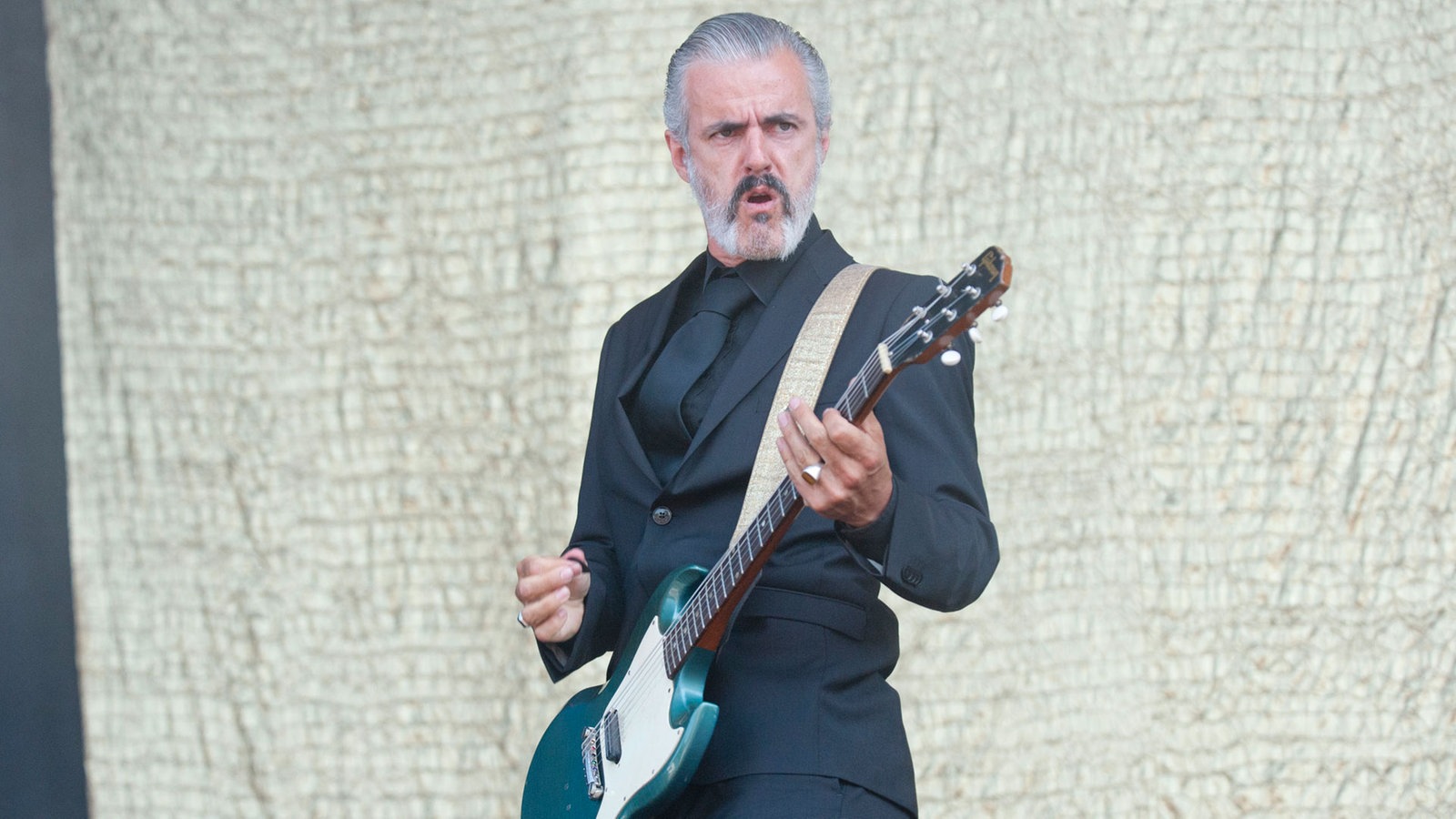Mann im schwarzen Anzug spielt Gitarre und schaut ernst zur Seite