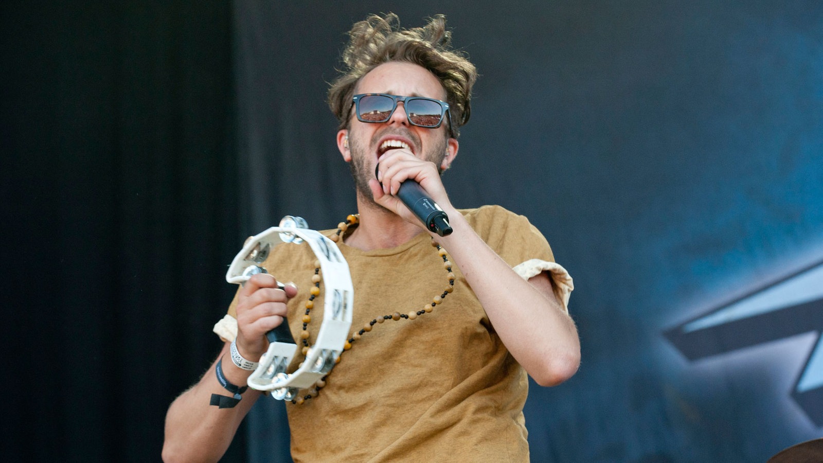 Sänger mit Sonnenbrille und Tamburin singt in ein Mikrofon.