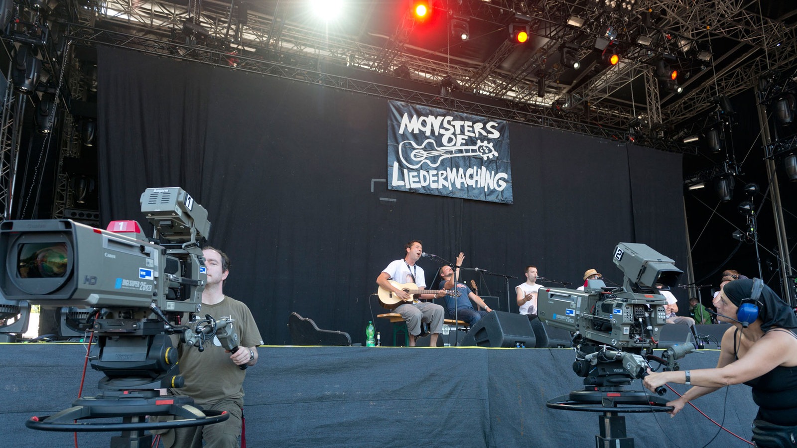 Kameras vor der Bühne, im Hintergrund spielt eine Band im Sitzen.