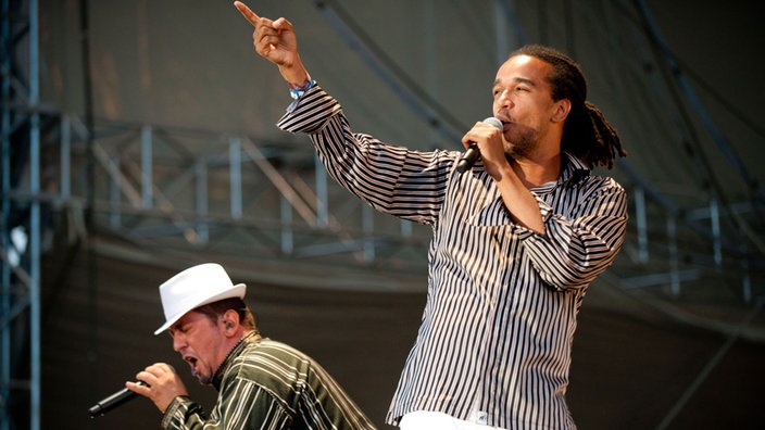 Hakim Meridja wird am Mikrofon von Aurélien Zohou unterstützt. Zohou trägt Dreadlocks und ein längs-gestreiftes hemd