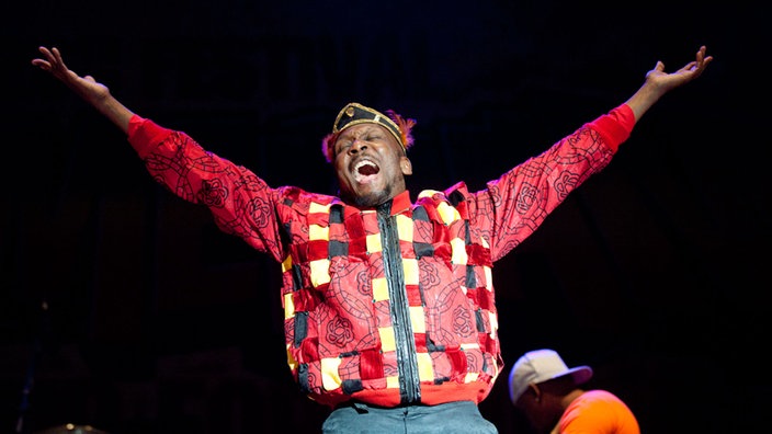 Jimmy Cliff hebt beide Arme in die Luft. Er trägt ein ausgefallen rot-kariertes Hemd und trägt eine Mütze dazu