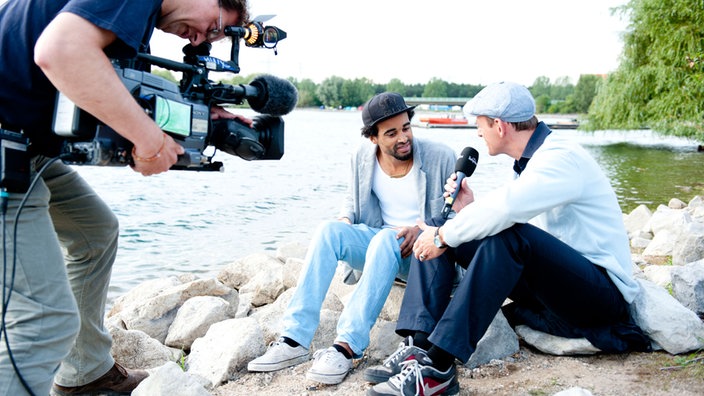 Patrice sitzt mit Interviewer Manuel Unger am See und wird dabei von einem Kameramann gefilmt.