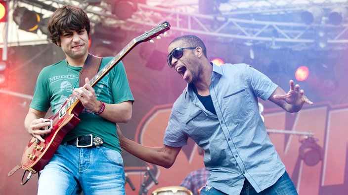 "Trombone Shorty" wird von einem Gitarristen im grünen T-Shirt begleitet. Shorty greift diesen aufmunternd an das T-Shirt.
