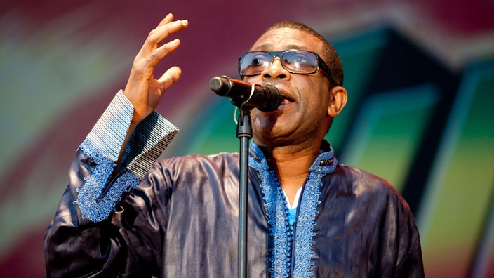 Youssou N'Dour hebt seinen linken Arm als wolle er Gott anrufen. Er trägt eine Sonnenbrille und blickt gen Himmel
